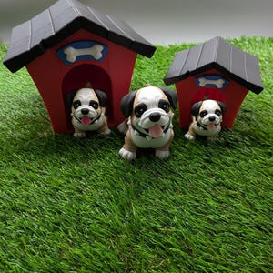 Op de afbeelding: Drie kleine bulldog-figuurtjes, &eacute;&eacute;n kijkt uit een rood hondenhok met een wit bot, en twee staan voor het hondenhok. Het hondenhok heeft een zwart dak en staat op een groene grasmat.