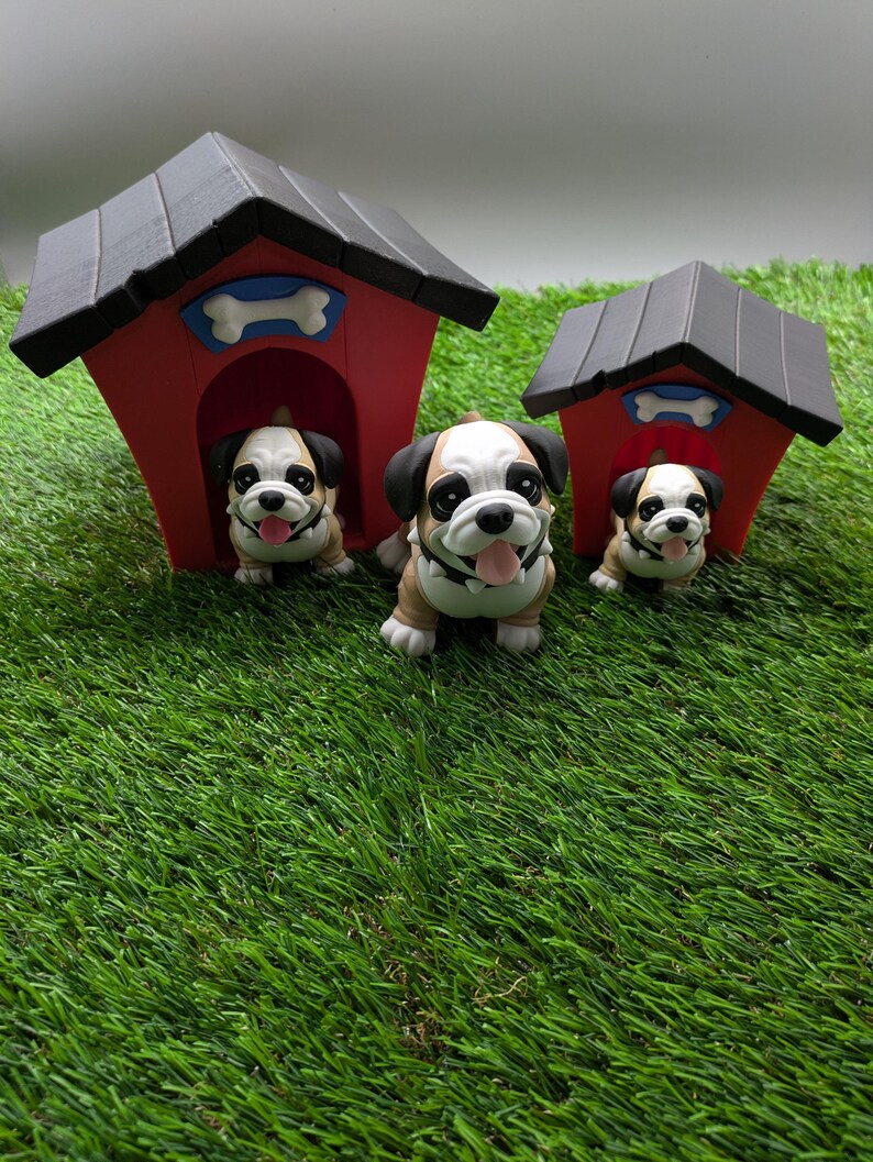 Op de afbeelding: Drie kleine bulldog-figuurtjes, &eacute;&eacute;n kijkt uit een rood hondenhok met een wit bot, en twee staan voor het hondenhok. Het hondenhok heeft een zwart dak en staat op een groene grasmat.