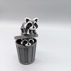 May include: A gray and white plastic figurine featuring two raccoons. One raccoon stands on top of a gray trash can lid, while a second raccoon head peeks out from inside the gray cylindrical trash can container below the lid.