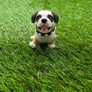 Op de afbeelding: Een klein, bruin en wit beeldje van een bulldog puppy met zwarte oren en neus. De puppy draagt een zwarte gespikkeld halsband en heeft een gelukkige uitdrukking op zijn gezicht. Het beeldje staat op een groene kunstgras ondergrond.