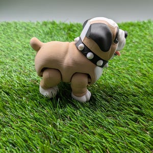 Op de afbeelding: Een bruine en witte bulldog-figuur met een zwarte neus en een gespikkeld kraag. De figuur staat op een groene kunstgras ondergrond.