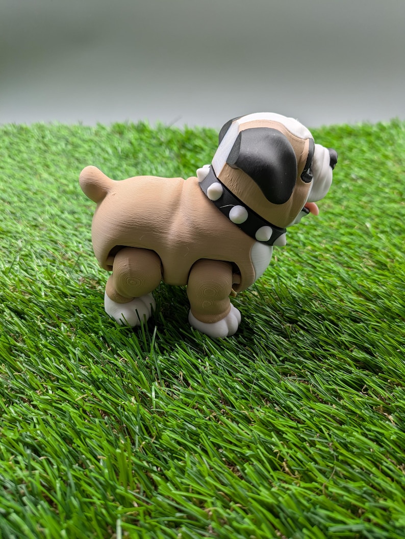Op de afbeelding: Een bruine en witte bulldog-figuur met een zwarte neus en een gespikkeld kraag. De figuur staat op een groene kunstgras ondergrond.