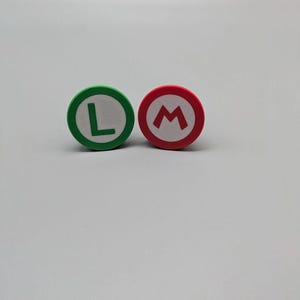 May include: Two round, colorful buttons. One is green with a white center and a green "L". The other is red with a white center and a red "M". These buttons are likely inspired by the popular video game series.