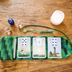 May include: A green and black crocheted tarot card holder with three cards displayed. The cards show illustrations and numbers. A blue stone, clear stones, and a white candle are also present on a wooden surface.
