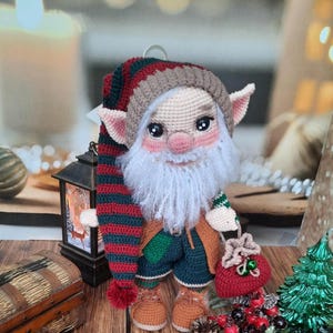 Könnte beinhalten: Gehäkelte Elfenpuppe mit einer rot-grünen gestreiften Mütze, braunen Stiefeln und einem grün-braunen Outfit. Der Elf hält eine rote Tasche mit einer grünen Glocke. Die Puppe sitzt auf einer Holzoberfläche mit einer Holzkiste, einer Laterne und Weihnachtsdekoration.