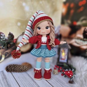 Könnte beinhalten: Eine gehäkelte Puppe mit einem rot-weiß gestreiften Hut, einer roten Jacke, einem blauen Rock und roten Schuhen. Die Puppe hat blonde Haare und steht auf einer Holzoberfläche.