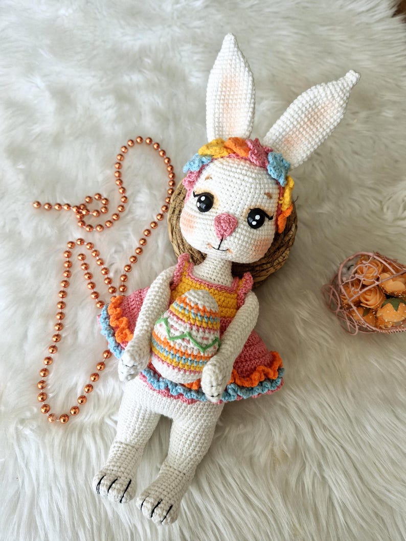 May include: A handmade white crochet bunny doll with large ears, wearing a pink and orange dress and a floral headband. The bunny holds a crochet Easter egg. A copper-colored beaded necklace is next to the bunny.