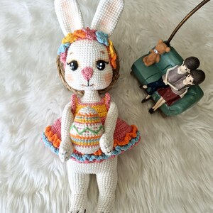 May include: A handmade crochet bunny doll with large white ears, wearing a colorful dress with an Easter egg design. The doll has a pink nose, big eyes, and a floral headband. The doll is on a white fluffy surface.