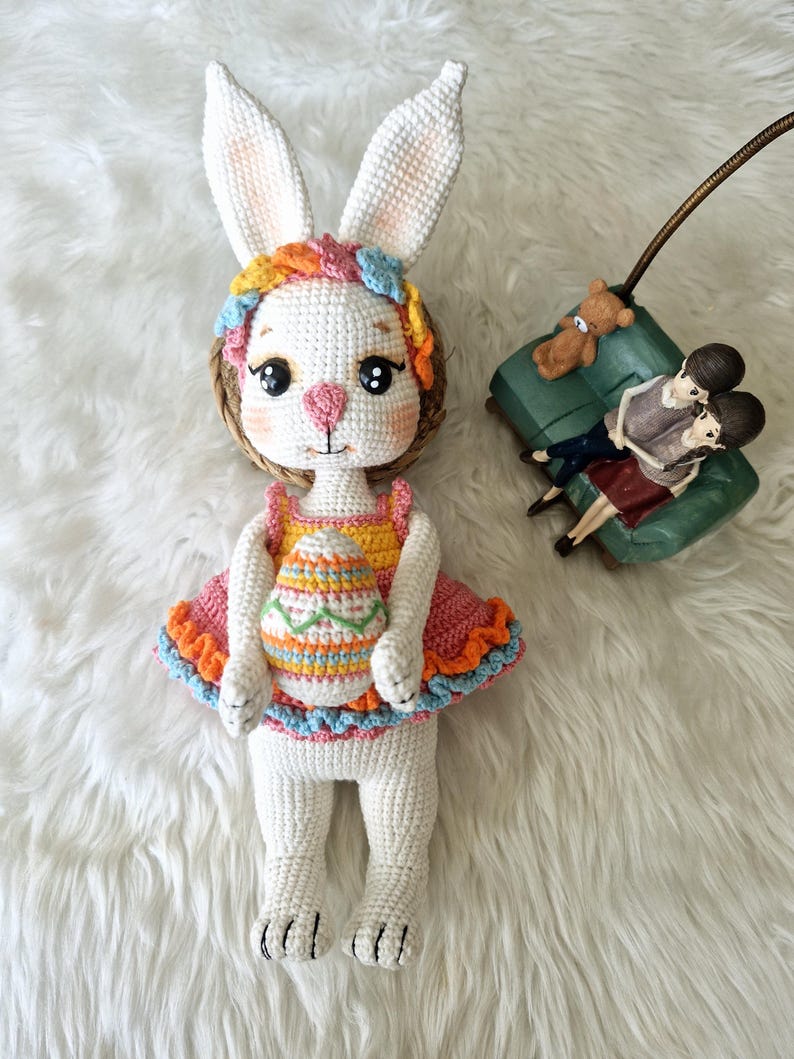 May include: A handmade crochet bunny doll with large white ears, wearing a colorful dress with an Easter egg design. The doll has a pink nose, big eyes, and a floral headband. The doll is on a white fluffy surface.