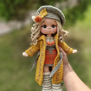 May include: A handmade crochet doll with blonde curly hair wearing a sage green beret adorned with a small orange pumpkin and yellow leaf. The doll wears a long mustard yellow coat over an orange skirt, white striped leg warmers, and orange shoes.