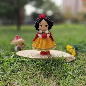 Op de afbeelding: Een handgemaakte gehaakte pop van Sneeuwwitje, gekleed in een gele jurk, rode cape en zwart haar met een rode strik. De pop houdt een rode appel vast en staat op een houten schijf in een grasveld.