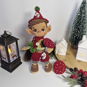 Peut inclure: Poupée elfe au crochet faite à la main, portant un chapeau, une écharpe et un short bordeaux. L'elfe a les cheveux bruns, la peau bronzée et de grandes oreilles. Il tient un champignon rouge et est décoré d'accents verts et blancs. La poupée mesure environ 20 cm de haut.