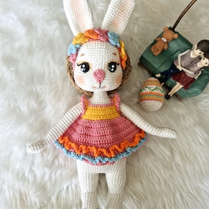 May include: A handmade crochet bunny doll with long ears, wearing a pink, yellow, and orange dress. The bunny has a colorful floral headband and rosy cheeks. An Easter egg and a small figurine are in the background.
