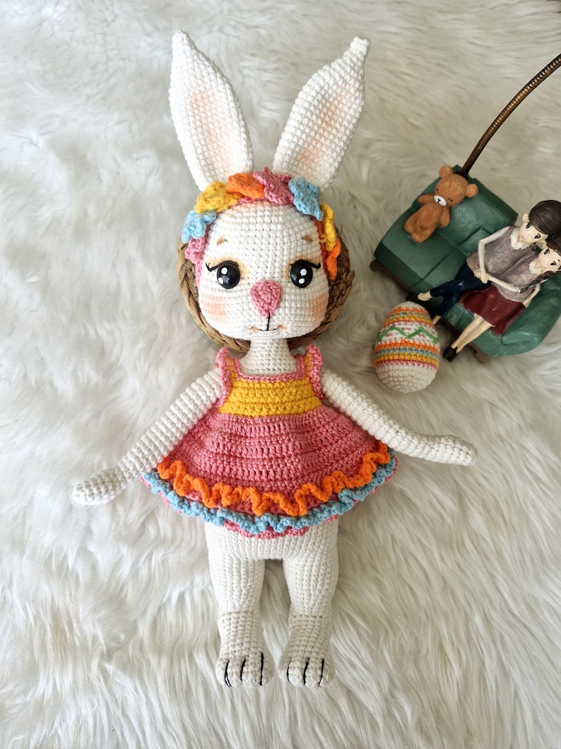 May include: A handmade crochet bunny doll with long ears, wearing a pink, yellow, and orange dress. The bunny has a colorful floral headband and rosy cheeks. An Easter egg and a small figurine are in the background.
