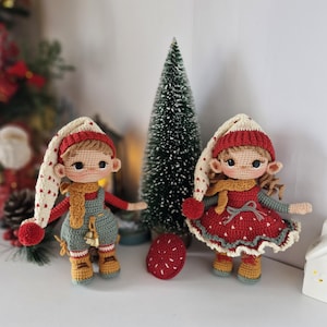 Op de afbeelding: Twee handgemaakte gehaakte poppen, één in een tuinbroek en de andere in een jurk, beide met lange, puntige hoeden. De poppen hebben een gebruinde huid, bruin haar en dragen rode, witte en gouden kleding. Op de achtergrond staat een kleine kerstboom.