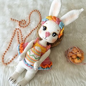 May include: A handmade white crochet bunny doll wearing a colorful dress and holding an Easter egg. The bunny has long ears, a floral headband, and is lying on a white fluffy surface. A beaded necklace and a small basket of flowers are also present.