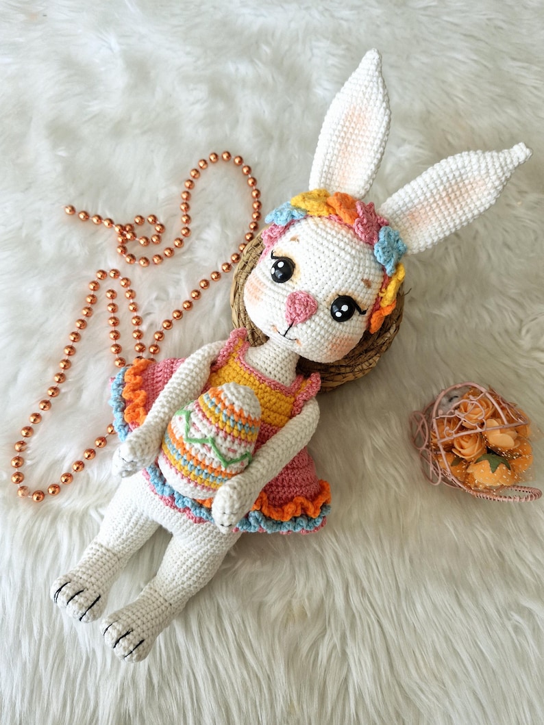 May include: A handmade white crochet bunny doll wearing a colorful dress and holding an Easter egg. The bunny has long ears, a floral headband, and is lying on a white fluffy surface. A beaded necklace and a small basket of flowers are also present.