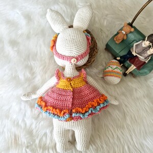 May include: A handmade crochet bunny doll with a pink and orange dress, blue trim, and a yellow top. The bunny has long ears and a pink headband. An Easter egg and a small green couch are in the background.