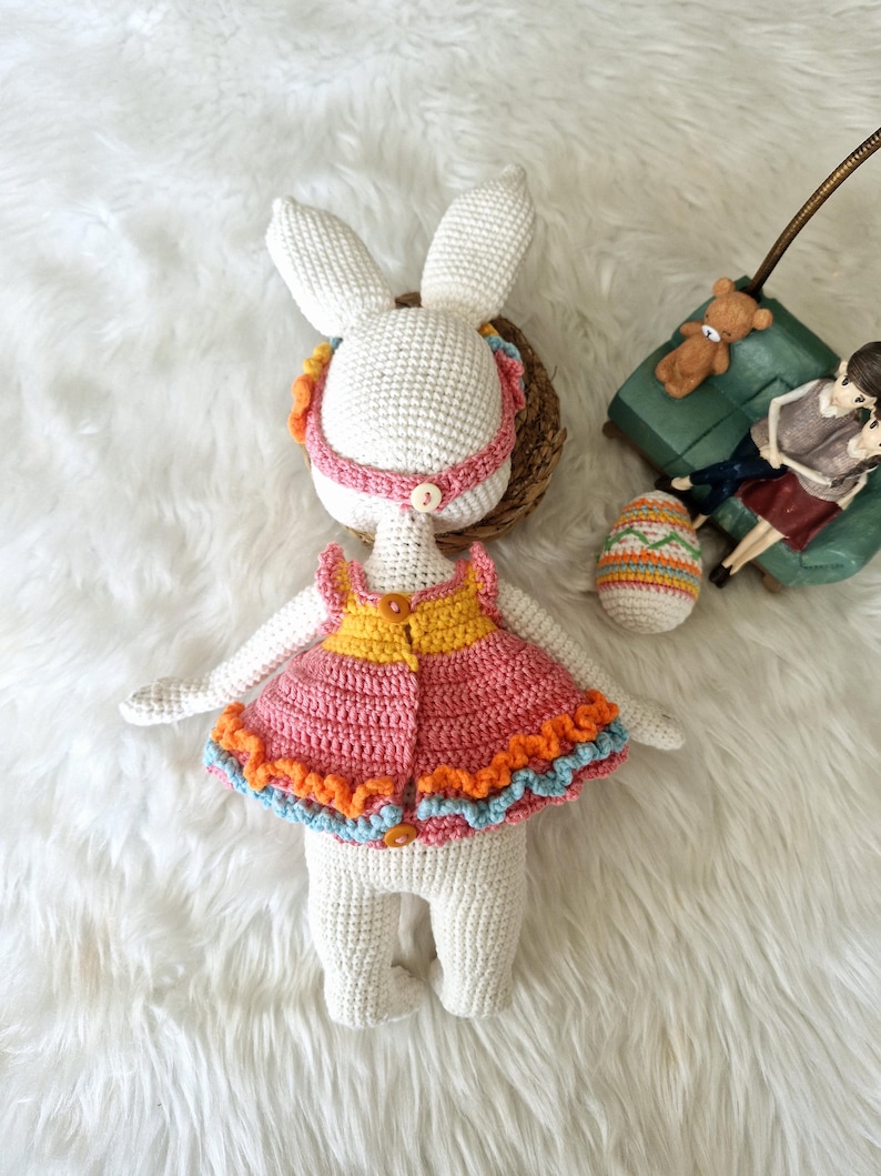 May include: A handmade crochet bunny doll with a pink and orange dress, blue trim, and a yellow top. The bunny has long ears and a pink headband. An Easter egg and a small green couch are in the background.