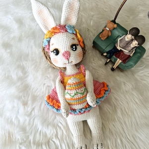 May include: A handmade white crochet bunny doll with long ears, wearing a colorful dress and holding an Easter egg. The doll has a pink nose, black eyes, and a floral headband. The dress is orange, pink, and blue.
