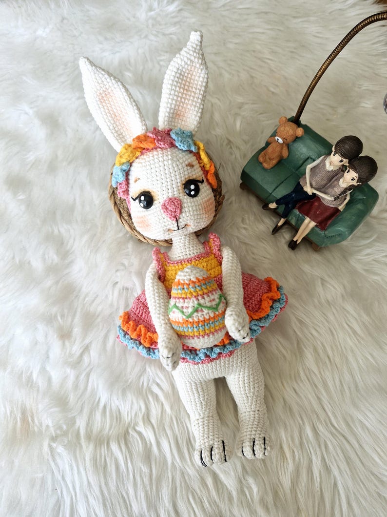 May include: A handmade white crochet bunny doll with long ears, wearing a colorful dress and holding an Easter egg. The doll has a pink nose, black eyes, and a floral headband. The dress is orange, pink, and blue.