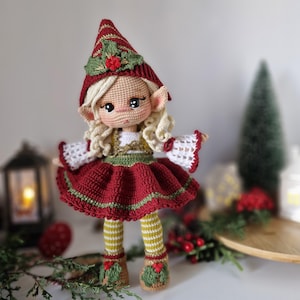 Può includere: Bambola elfo all'uncinetto fatta a mano. La bambola ha capelli biondi ricci, guance rosee e indossa un vestito rosso con finiture verdi, un cappello abbinato e calze a righe. Le scarpe dell'elfo sono decorate con agrifoglio.