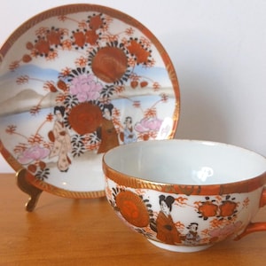 Peut inclure: Une tasse à thé et une soucoupe en porcelaine blanche avec un motif d'inspiration japonaise. La tasse et la soucoupe présentent un motif floral avec des accents dorés et une représentation de personnages en vêtements japonais traditionnels.