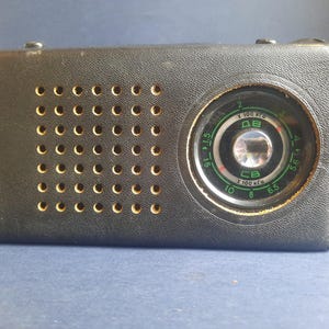 Peut inclure: Un radio transistor vintage noir avec un cadran rond et une grille de haut-parleur. Le cadran a une échelle verte avec des chiffres et le texte "x 100 KHz" et "x 100 MHz".