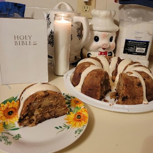 May include: A slice of cake on a floral plate, next to a whole cake on a white plate. A white Bible, a lit candle, and a pig-shaped container are in the background. The cake has a white glaze.