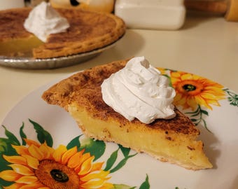 May include: A slice of pie with a golden-brown crust and creamy yellow filling, topped with a dollop of whipped cream. The pie sits on a plate decorated with sunflowers and green leaves. Another pie is in the background.