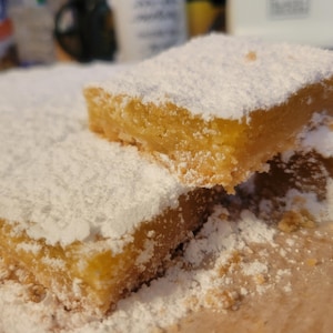 May include: Two square lemon bars with a golden yellow citrus filling and a shortbread crust, generously dusted with a thick layer of white powdered sugar. The bars are arranged with one piece resting partially on top of the other piece.