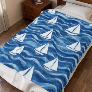 May include: A blue and white blanket featuring a nautical design. The blanket displays white sailboats sailing on blue watercolor waves. The blanket is on a bed with a white pillow and a wooden nightstand.