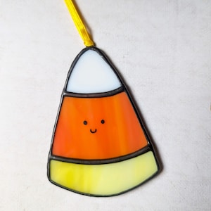 Peut inclure: Ornement en vitrail en forme de bonbon de maïs avec un visage souriant. L'ornement présente des sections blanches, orange et jaunes, soulignées de noir. Un ruban jaune est attaché pour l'accrocher. Décoration festive pour Halloween.