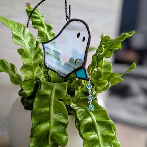 May include: A stained glass ghost decoration with a black wire frame, two black dot eyes, and a turquoise bow. The ghost hangs from a black wire loop and is adorned with three blue crystal beads. The ghost is set against a backdrop of green plants.