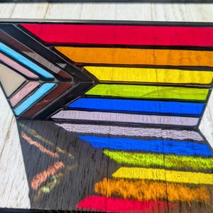 Peut inclure: Pièce d'art en vitrail représentant le drapeau Progress Pride. L'œuvre présente un éventail de couleurs vives, dont le rouge, l'orange, le jaune, le vert, le bleu, le violet, le rose, le bleu clair, le marron et le blanc, dans un cadre noir. Suspendu par une chaîne.