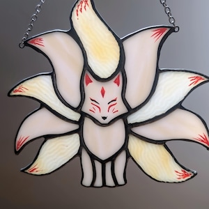 May include: Stained glass fox with nine tails, cream-coloured body and tails with yellow and red accents. The fox has a red face with detailed features and is outlined in black. A silver chain is attached for hanging.