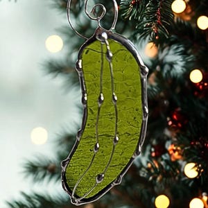 Peut inclure: Ornement de Noël en vitrail en forme de cornichon. Le cornichon est vert translucide texturé avec des accents argentés et une boucle en fil d'argent pour l'accrocher. L'ornement est suspendu devant un sapin de Noël flou.
