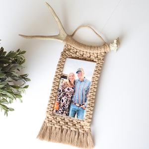 Puede incluir: Un marco de fotos de macramé con un acento de cuerno natural. El marco está hecho de cuerda beige tejida y presenta una abertura rectangular para una fotografía. La parte inferior del marco tiene un detalle de flecos. La foto muestra una pareja sonriendo.