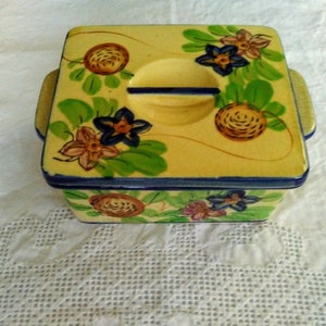 May include: A yellow ceramic square dish with a lid. The lid has a floral design with blue, pink, and green flowers. The dish has a floral design with brown, pink, and green flowers.
