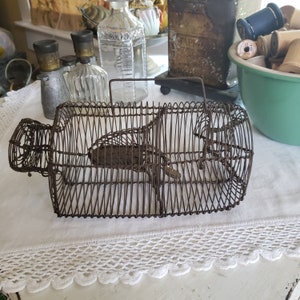 May include: A rusty metal cage trap for catching small animals. The trap is made of wire and has a hinged door. The trap is sitting on a white tablecloth with lace trim.