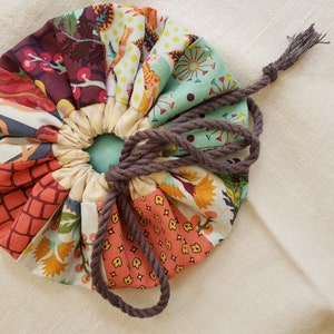 May include: A round, drawstring bag made of patchwork fabric with a variety of colorful floral and geometric patterns. The bag is tied closed with a brown cord.