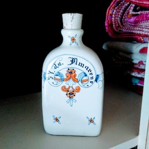 May include: White ceramic bottle with a cork stopper. The bottle is decorated with blue, orange, and floral designs, and the text "I. de. Amarre" is written on the front. The bottle is rectangular with rounded edges.