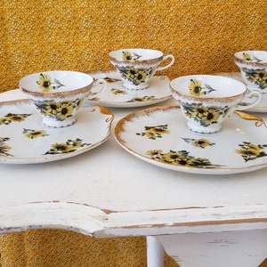 May include: A set of four white teacups and saucers with a sunflower design. The cups and saucers have gold trim. The saucers are shaped to hold the cups and have a decorative edge.