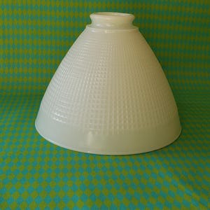 May include: A white, cone-shaped lampshade with a textured, grid-like pattern. The lampshade sits on a teal and green diamond-patterned surface. The lampshade has a wide base and a narrow top opening.