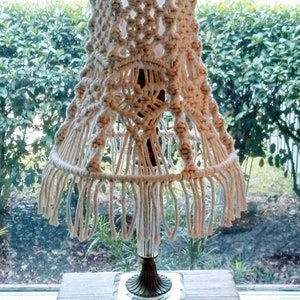 May include: A white macrame lampshade with fringe hangs from a clear glass lamp base with a gold metal accent. The lamp is sitting on a wooden table.
