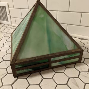 May include: A pyramid-shaped stained glass lamp shade with a bronze-colored metal frame. The glass is a mix of green and white, creating a marbled effect. The shade is sitting on a white and gray hexagonal tile surface.