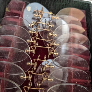 Pu&ograve; includere: Una collezione di occhiali pince-nez antichi con montature color oro e lenti trasparenti, disposti in una scatola nera con interno in velluto rosso. Gli occhiali sono di forma ovale e presentano un ponte per il naso.