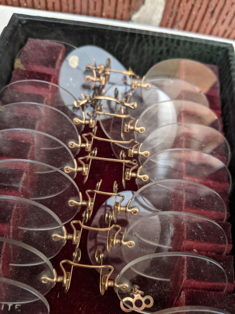 Pu&ograve; includere: Una collezione di occhiali pince-nez antichi con montature color oro e lenti trasparenti, disposti in una scatola nera con interno in velluto rosso. Gli occhiali sono di forma ovale e presentano un ponte per il naso.