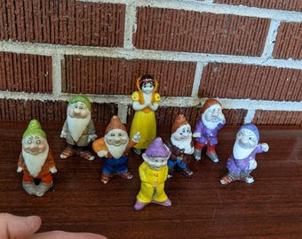 May include: A collection of ceramic figurines featuring Snow White and the Seven Dwarfs. The figurines are brightly colored, with details like hats, beards, and clothing. The figures are arranged on a dark brown surface.