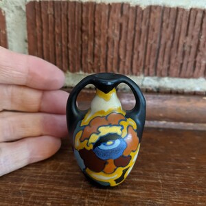 May include: A small, decorative ceramic vase with a black top and handles. The vase features a colorful floral design in shades of blue, brown, yellow, and orange. The vase is approximately 3 inches tall.